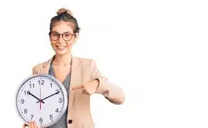 Beautiful caucasian woman with blonde hair wearing business jacket and holding clock smiling happy pointing with hand and finger